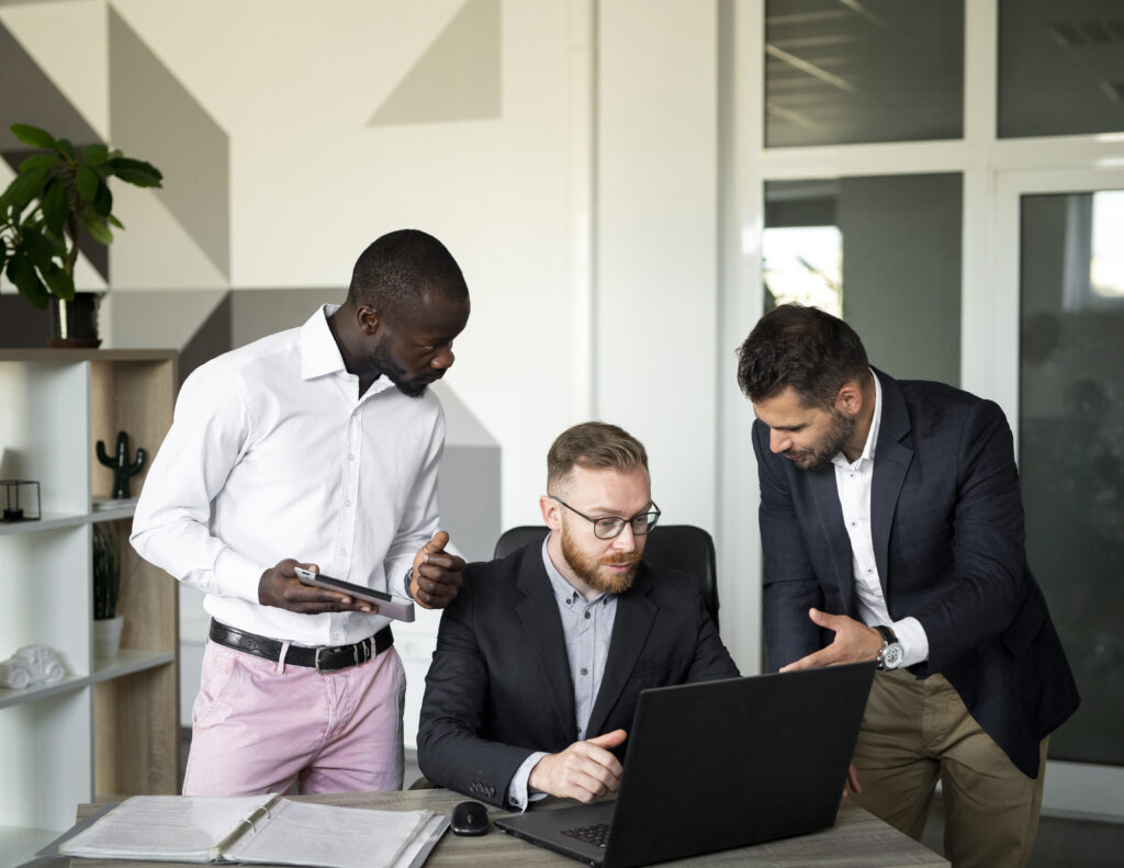 A man sitting staring at his laptop while two men standing while they try to explain to the man