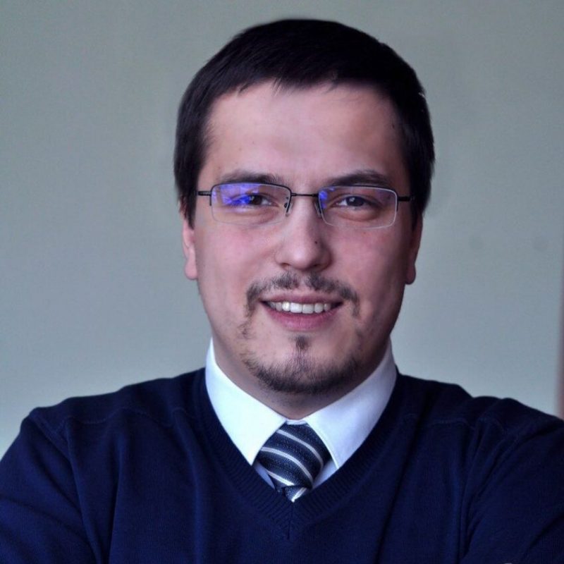 Alexander Shalkin, Canada Digital Advisor and CEO, dons a blue sweater over a white shirt with a tie, complemented by glasses, as he confidently faces the camera.