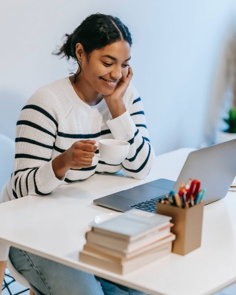A woman smiling holding a mug while staring at a laptop screen